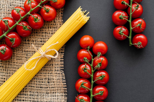 Top view of raw pasta and cherry tomatoes on sackcloth on black background  - Valokuva, kuva