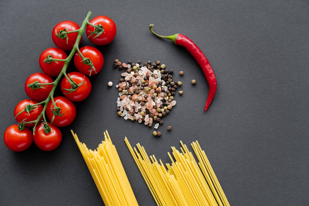 Draufsicht auf ungekochte Pasta in der Nähe von Gewürzen und frischem Gemüse auf schwarzem Hintergrund  - Foto, Bild