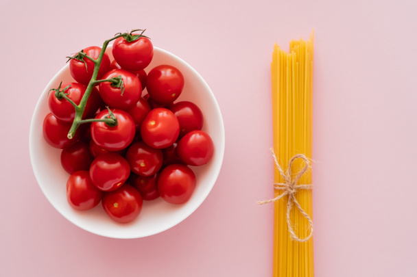 Top view of cherry tomatoes in bowl and uncooked spaghetti on pink surface - Photo, image