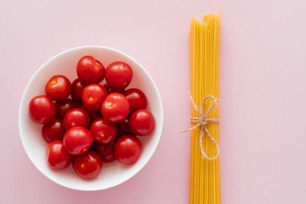 Vista superior de tomates cherry frescos en tazón y espaguetis sin cocer en la superficie rosa - Foto, Imagen