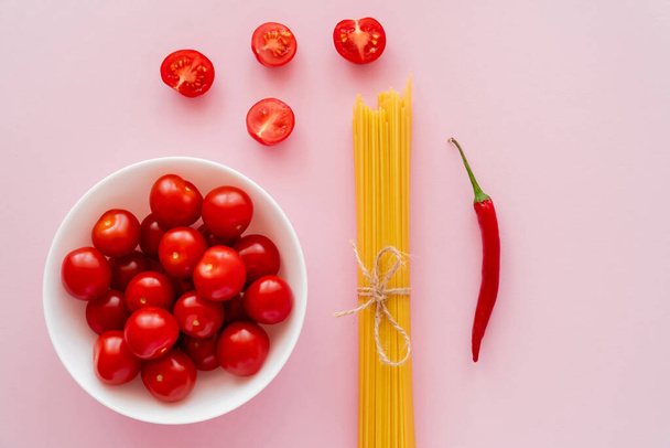 Top view of cherry tomatoes in bowl near raw pasta and chili pepper on pink surface - 写真・画像