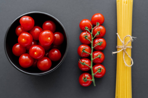 Vista superior de tomates cherry en un tazón cerca de espaguetis sin cocer en la superficie negra  - Foto, Imagen