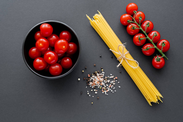 Top view of cherry tomatoes in bowl near raw pasta and spices on black surface  - Fotó, kép