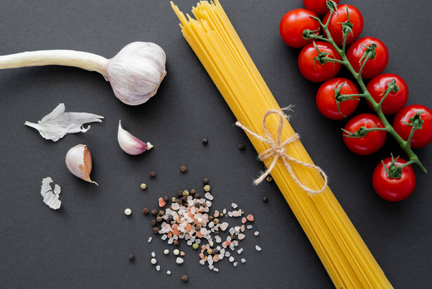Top view of garlic and cherry tomatoes near spices and raw pasta on black surface  - Foto, Imagem