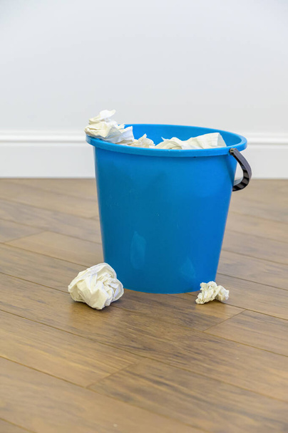 a blue plastic bucket filled with used tissues sits on a wooden floor. - Zdjęcie, obraz