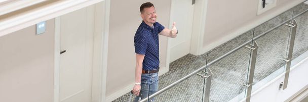 Portrait of cheerful tourist man with suitcase search for his room in hotel show thumbs up gesture. Male come for holiday or business trip. Travel concept - Фото, зображення