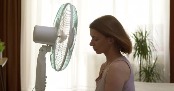 Una mujer se sienta cerca de un ventilador giratorio en una corriente de aire fresco en una habitación caliente en verano. - Metraje, vídeo