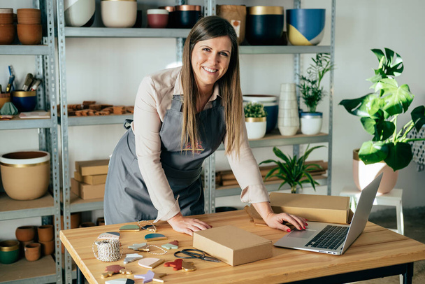 Portrait Of Smiling Woman In Apron Using A Laptop Computer And Preparing Christmas Ornaments Of Clay For Delivery At Her Store - Φωτογραφία, εικόνα