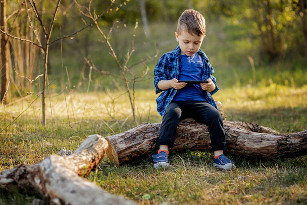 Cheerful child sitting on a fallen tree, watching cartoons on the phone in the summer. Cute boy having fun in nature. - Zdjęcie, obraz