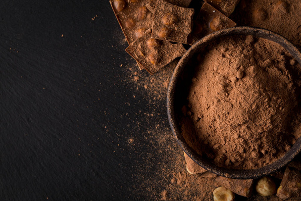 Broken chocolate nuts pieces and cocoa powder in bowl on dark stone background - Photo, image