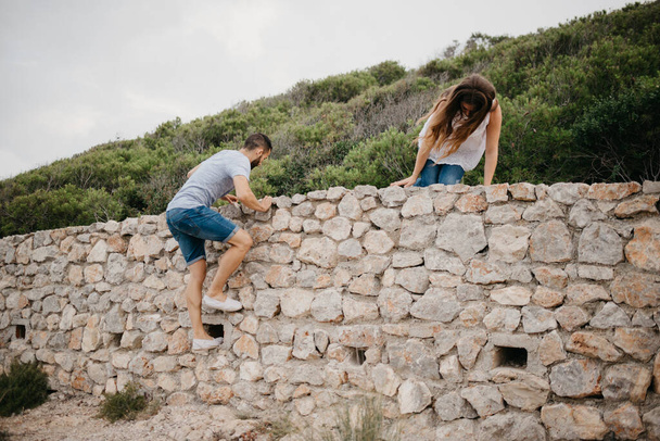 Ένας Ισπανός και η κοπέλα του σκαρφαλώνουν πάνω από τον αρχαίο πέτρινο τοίχο σε ένα πάρκο στην Ισπανία. Μερικοί τουρίστες απολαμβάνουν την πεζοπορία μεταξύ τους σε μια ημερομηνία στο ηλιοβασίλεμα στη Βαλένθια. - Φωτογραφία, εικόνα