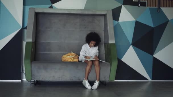 Komoly tinédzser lány, aki a házi feladatát csinálja a modern iskolateremben egyedül. Koncentrált göndör afro-amerikai lány ül padon élénk folyosón. Gondolatos kreatív gyermek rajz vázlatok notebook. - Felvétel, videó