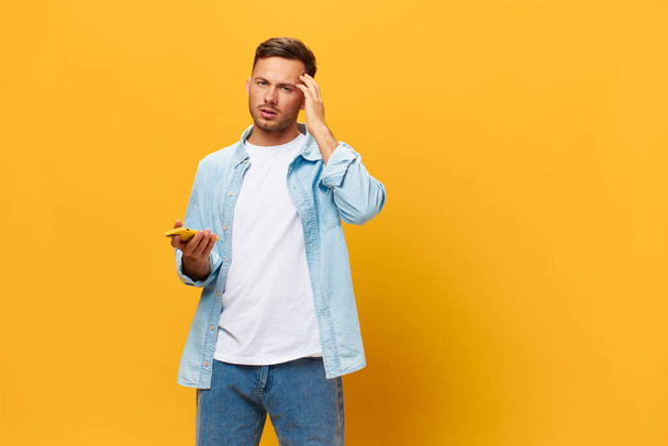 Thoughtful unhappy pensive handsome man in blue basic t-shirt make difficult decision hold phone posing isolated on yellow studio background. Copy space Banner Mockup. Online Social media concept - Photo, Image