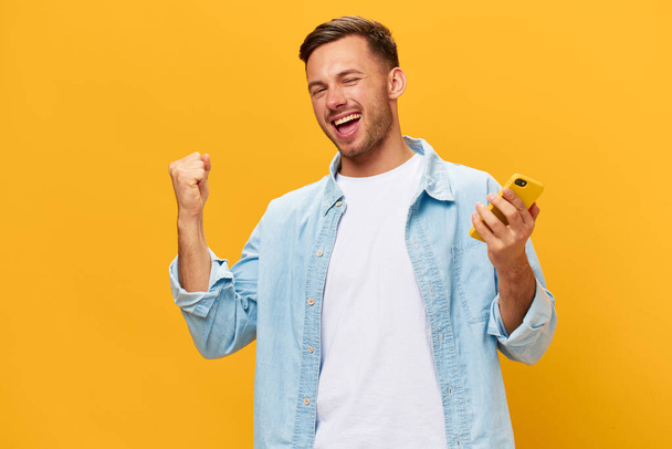 Iloinen ruskettunut komea mies sinisessä t-paidassa nostaa nyrkki ylös puhelimella poseeraa eristetty oranssi keltainen studio tausta. Kopioi tilaa Banner Mockup. Online sosiaalinen media. Lucky win käsite - Valokuva, kuva