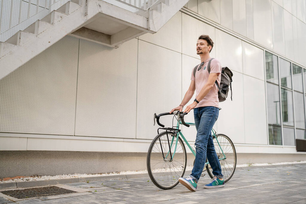 One man young adult male with brown hair and mustaches walking by the building with rucksack on his back and bicycle happy smile joyful real people copy space side view full length - Фото, изображение