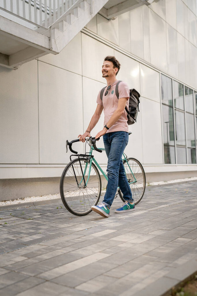 One man young adult male with brown hair and mustaches walking by the building with rucksack on his back and bicycle happy smile joyful real people copy space side view full length - Фото, зображення