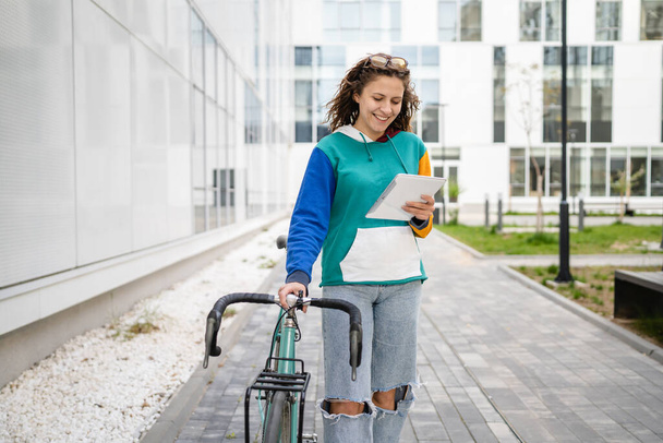 Yetişkin bir kadın, elinde bisiklet ve dijital tabletle üniversite binasının önünden geçiyor. Gerçek insanlar uzay gülüşünü neşeli bir şekilde kopyalıyor. - Fotoğraf, Görsel