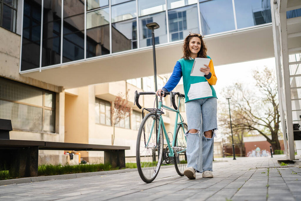 Одна жінка молода доросла біла жінка, що йде університетською будівлею, тримає велосипед та цифровий планшет удень, справжні люди копіюють простір щасливим повнометражним видом спереду
 - Фото, зображення