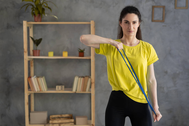 portrait of sporty woman workout with rubber resistance band, performs exercises for the muscles of back and hands. Indoor home workout concept - Φωτογραφία, εικόνα