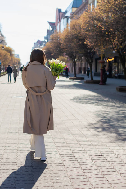 Back view of girl in white costume and beige coat walking on the street and holding bouquet of white fresh spring tulip flowers. Concept of fashion clothes, spring or gift for the March 8 womens day. - Photo, Image