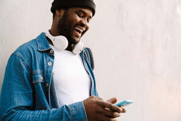 Black young man with headphones smiling and using mobile phone outdoors - Foto, Imagem