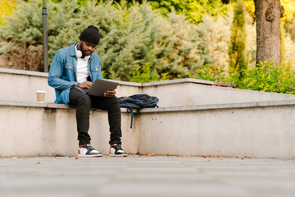 Black man with headphones working with laptop while sitting on bench outdoors - Φωτογραφία, εικόνα