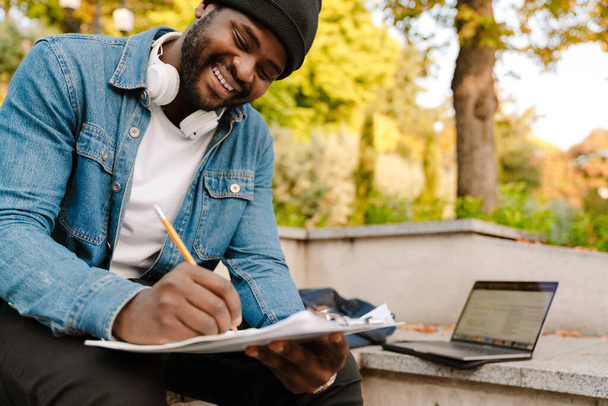 Black man working with laptop and papers while sitting on bench in park - Foto, immagini