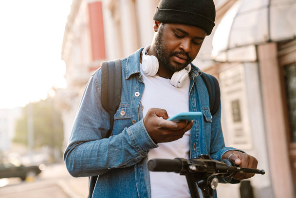 Black man with headphones using mobile phone while riding scooter outdoors - Foto, Imagem
