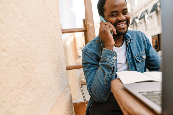 Černoch mluví na mobilu při práci s notebookem v kavárně venku - Fotografie, Obrázek