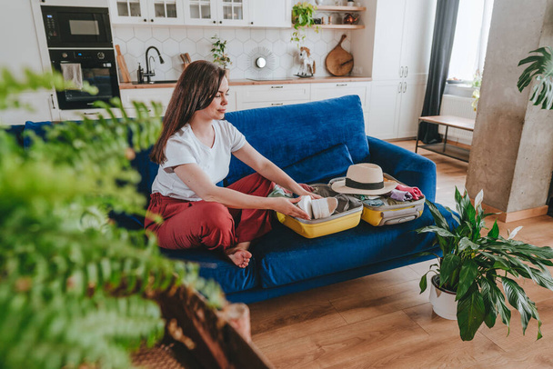 Beautiful smiling young woman packing personal belongings in a suitcase for travelling looking at list at tablet sitting on the sofa in the living room. - Foto, Imagen