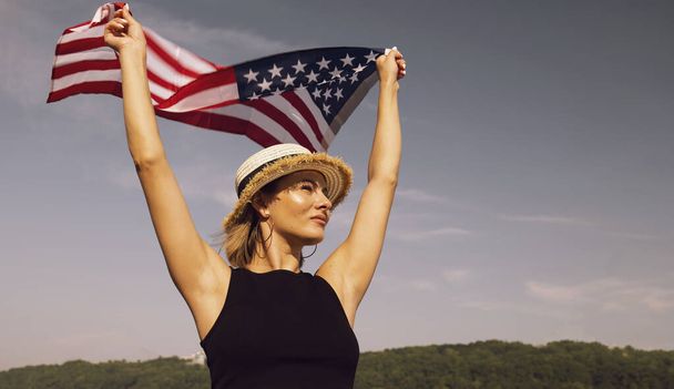 Młoda kobieta w kapeluszu z flagą USA na tle błękitnego nieba. Dzień FLAG. Urlop amerykański - Zdjęcie, obraz