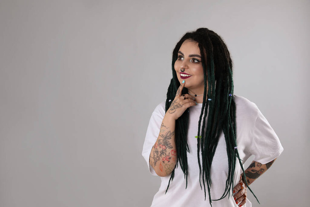 Beautiful young woman with tattoos on arms, nose piercing and dreadlocks against grey background. Space for text - Zdjęcie, obraz