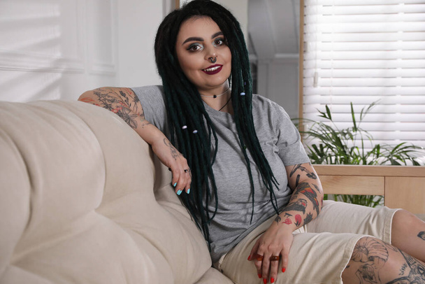 Beautiful young woman with tattoos on body, nose piercing and dreadlocks at home - Photo, image