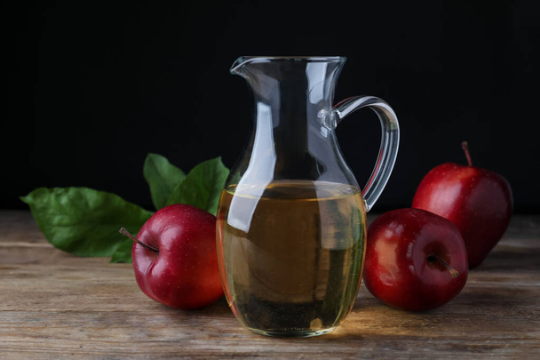 Jug of tasty juice and fresh red apples on wooden table against black background - Valokuva, kuva