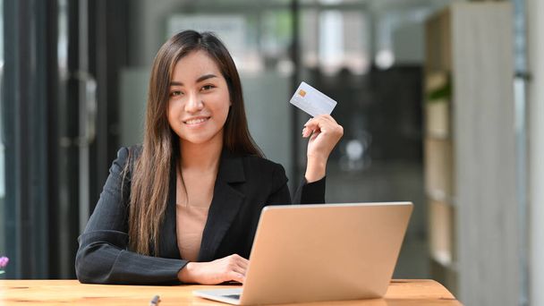 Confident asian woman sitting front of laptop and holding credit card in her hand. Online shopping, e-commerce concept. - Photo, Image
