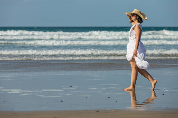 Side näkymä paljain jaloin naisten turisti valkoinen mekko koskettaa olki hattu ja tarkkailemalla rannalla kävellessään märkä hiekka lähellä vilkuttaa merta aurinkoisena kesäpäivänä - Valokuva, kuva