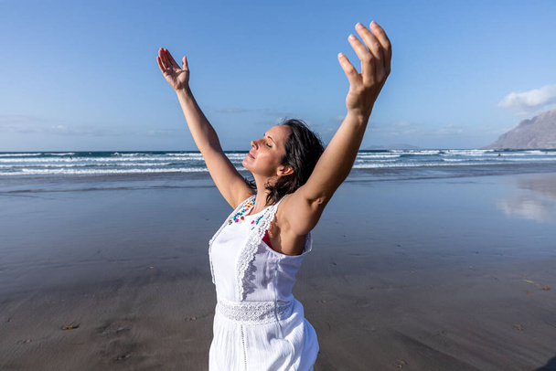 リゾートの晴れた夏の日に波の海に近い湿ったビーチで休んでいる間、中年の女性旅行者は腕を上げ、閉じた目で新鮮な空気を呼吸します - 写真・画像