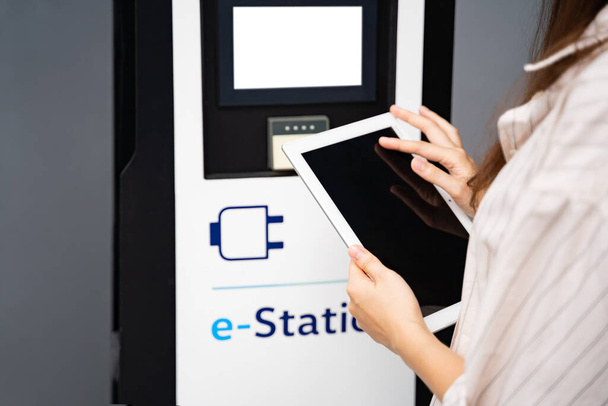 Young Asian or Caucasian woman wearing shirt holding and using white tablet, connecting to bluetooth touchscreen electric car charge station close up. Carsharing, modern technology, alternative energy - Fotoğraf, Görsel