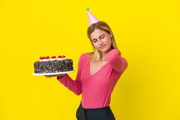 Sarışın Uruguaylı kız, sarı arka planda izole edilmiş doğum günü pastası tutuyor. Çabaladığı için omuz ağrısı çekiyor. - Fotoğraf, Görsel