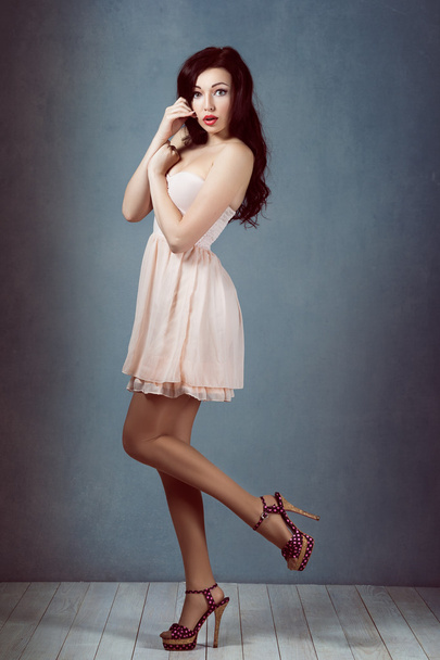 Sexy jeune belle brune aux cheveux longs, avec une silhouette élancée debout sur une jambe levante tenant son talon dans une robe rose et rétro femme sportive et acrobatique pin up maquillage
 - Photo, image