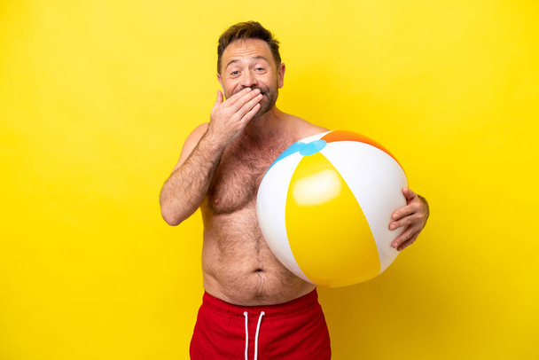 Homem caucasiano de meia idade segurando bola de praia isolada no fundo amarelo feliz e sorrindo cobrindo a boca com a mão - Foto, Imagem