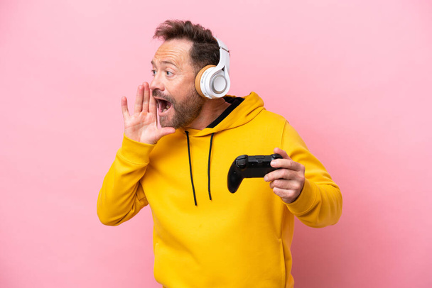 Uomo di mezza età che gioca con un controller di videogiochi isolato su sfondo rosa gridando con la bocca spalancata sul lato - Foto, immagini