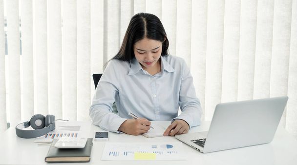 Azjatka pracująca na laptopie. Uśmiechnięta bizneswoman pracująca i notująca w biurze. - Zdjęcie, obraz