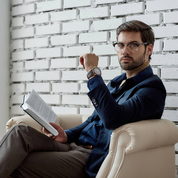 Caucasian IT businessman looking at camera while reading book in armchair in office. Business strategy, development, promotion and selling of high-tech company. Modern successful male lifestyle - 写真・画像