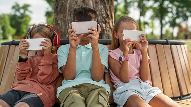 Obscure faces of children using and watching smartphones on wooden bench in park outdoors. Boys and girl of generation alpha. Gadget addiction. Childhood lifestyle. Sunny summer day - 写真・画像
