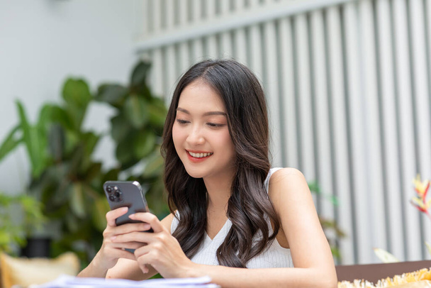Heureuse jeune femme asiatique sourire en utilisant smartphone surfer sur Internet et profiter avec les médias sociaux ou achats en ligne à la maison. Vie de bien-être moderne de mignonne femme regardant le plaisir et le bonheur de téléphone mobile - Photo, image