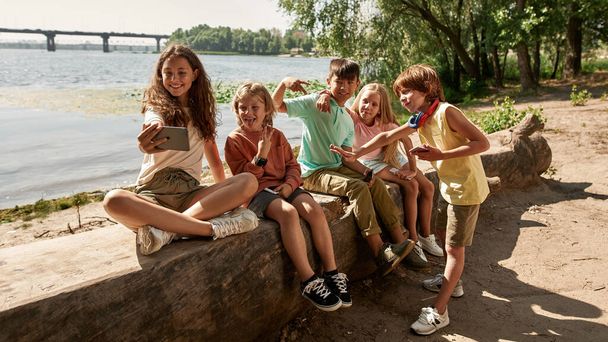 Group of smiling multiethnic children taking selfie on mobile phone on river coast in park. Boys and girls of generation alpha. Gadget addiction. Friendship. Childhood lifestyle. Warm summer day - Zdjęcie, obraz