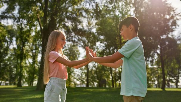 Side view of blonde girl and multiracial boy play patty cake game on green meadow in blurred park outdoors. Smiling caucasian children of generation alpha. Childhood lifestyle. Friendship. Sunny day - Foto, Imagen