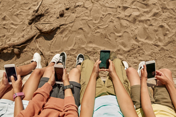 Cropped top view of group of children using and browsing smartphones on log on sandy coast outdoors. Boys and girl of generation alpha. Gadget addiction. Childhood lifestyle. Sunny summer day - Fotografie, Obrázek