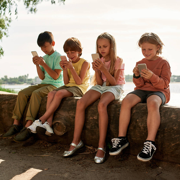 Group of multiethnic children using and browsing smartphones on log on river coast outdoors. Boys and girl of generation alpha. Gadget addiction. Childhood lifestyle. Sunny summer day - Foto, imagen